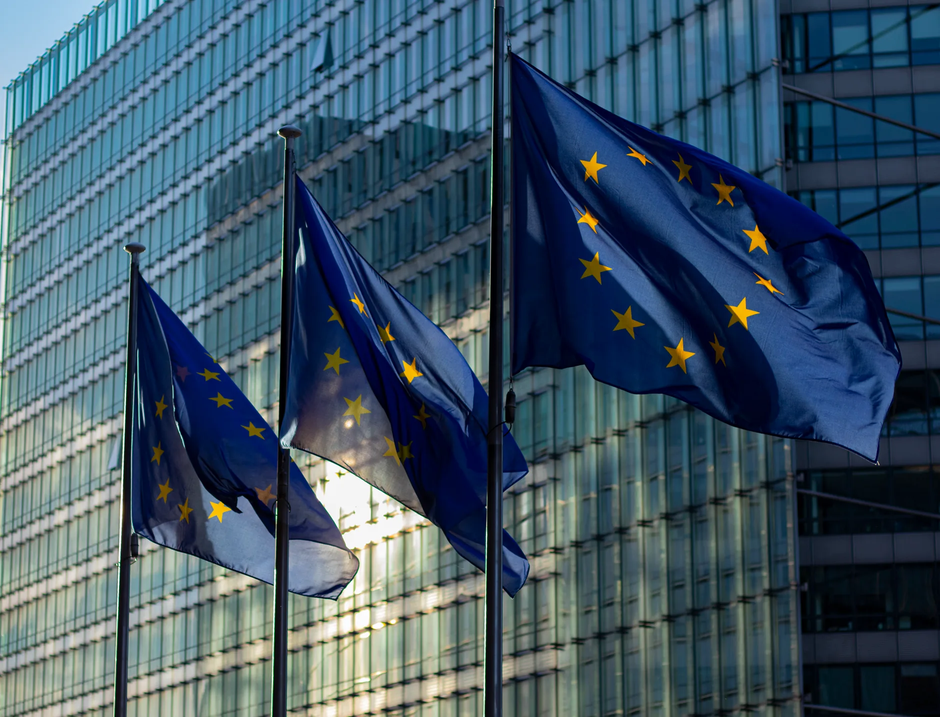 Trois drapeaux européens flottent devant la façade vitrée d'un bâtiment institutionnel