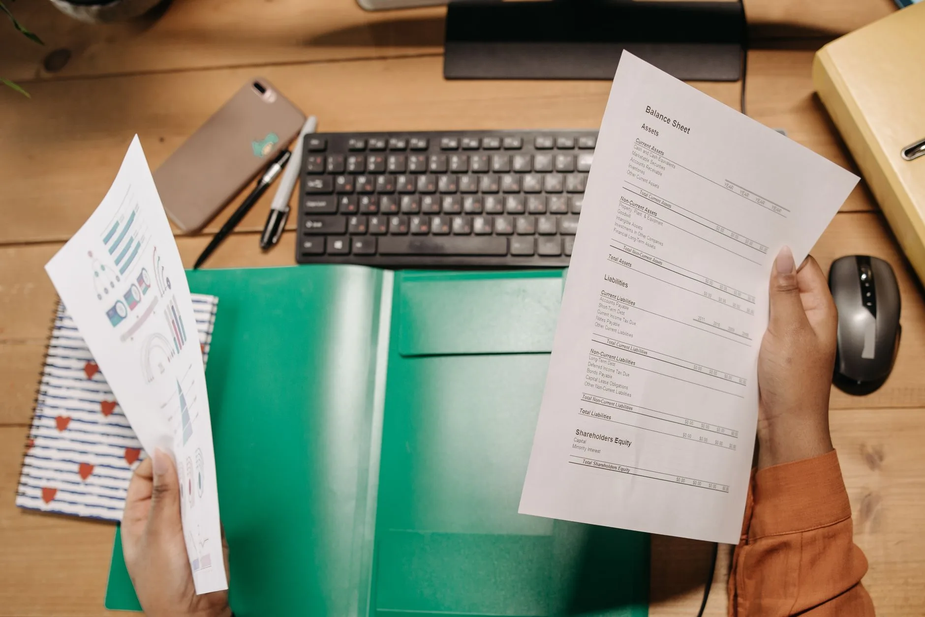 Deux mains comparent des factures papier au-dessus d'un clavier et d'un classeur vert