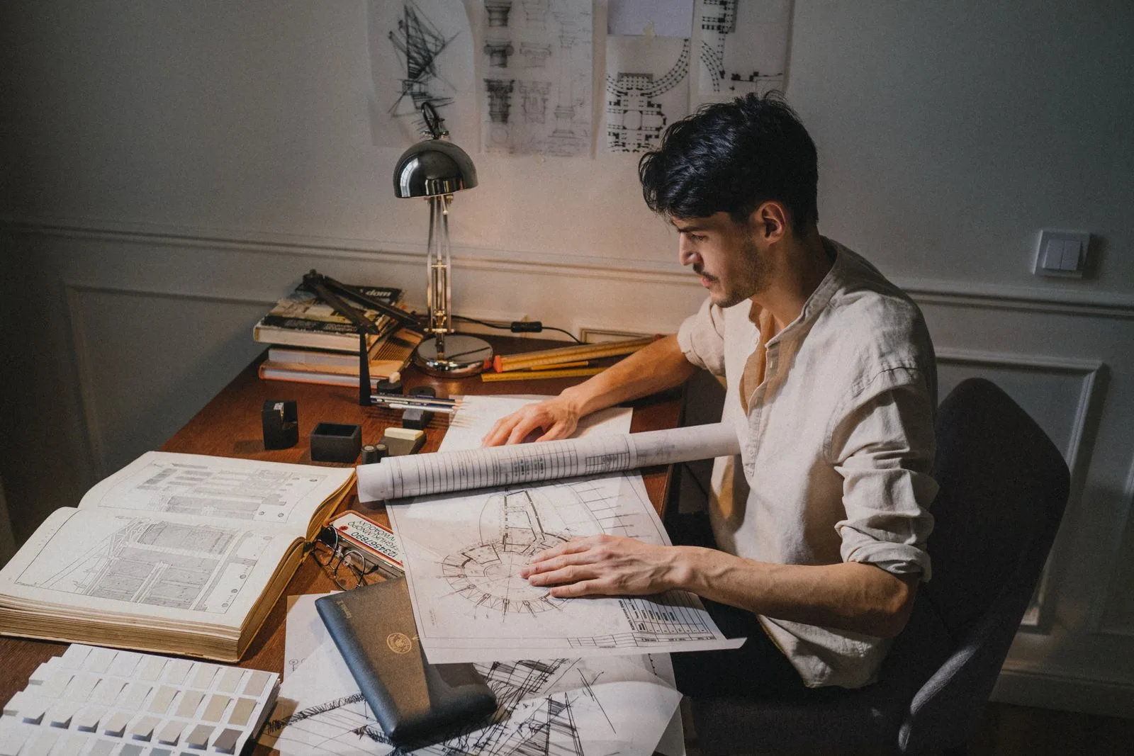 Projeteur qui consulte un plan roulé et un ouvrage de référence à son bureau, sous une lampe