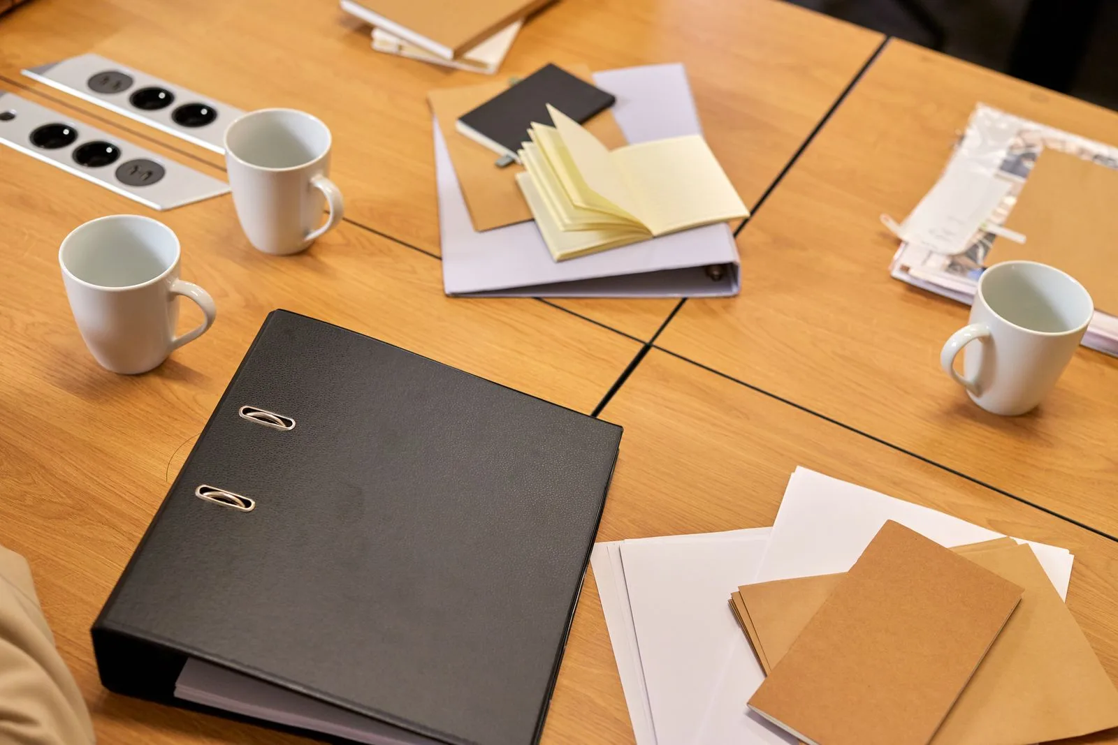 Classeur noir, carnets et tasses posés sur une table de réunion en bois
